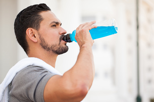 Closeup of a handsome young man with a beard drinking a sports drink from a bottle after running outdoors in the city