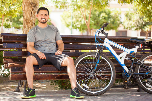 Full length portrait of a handsome young man sitting on a park bench after taking a bike ride around the city