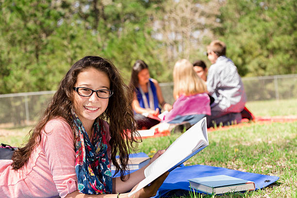 Education: Latin pre-teenage girl studies at local park. Friends background. Pretty, Latin descent teenage girl studies or reads a book at a local park or school campus. She holds an open notebook. Multi-ethnic group of friends in background. 13 14 years child teenage girls lying down stock pictures, royalty-free photos & images