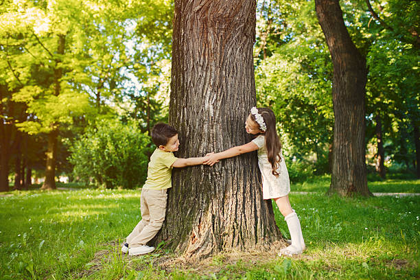 Brother and sister are playing in the park holding hands and hugging tree.