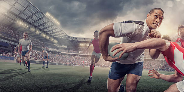 Rugby Player Running With Ball Whilst Being Tackled During Match A close up image of a professional male rugby player running with a rugby ball under his arm through an opposition player trying to tackle him. The action takes place during a rugby game in a generic outdoor floodlit rugby stadium full of spectators under a dramatic stormy sky.  rugby team stock pictures, royalty-free photos & images