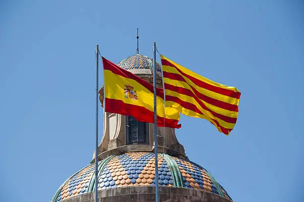 Spanish and Catalan flag flying together against blue sky on a bank building in Barcelona, Spain