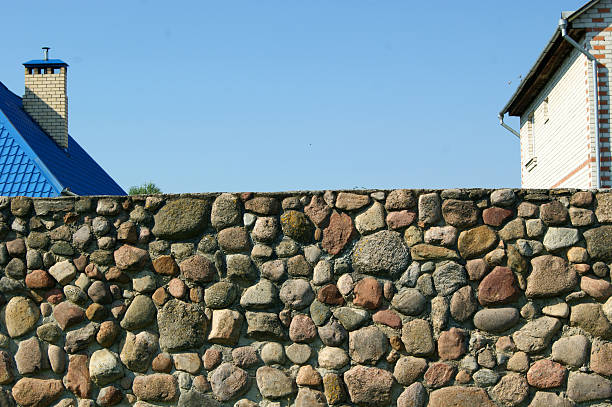 Konevets Island, Russia, July 13, 2024. Rocky area with large stones and purple wildflowers near a sandy path. Three people walk on the path, dressed casually one wears a headscarf. Another person, also with a headscarf, sits on the rocks. Surrounding area features dense green trees. Bright sunlight illuminates the scene, creating a natural, tranquil setting.