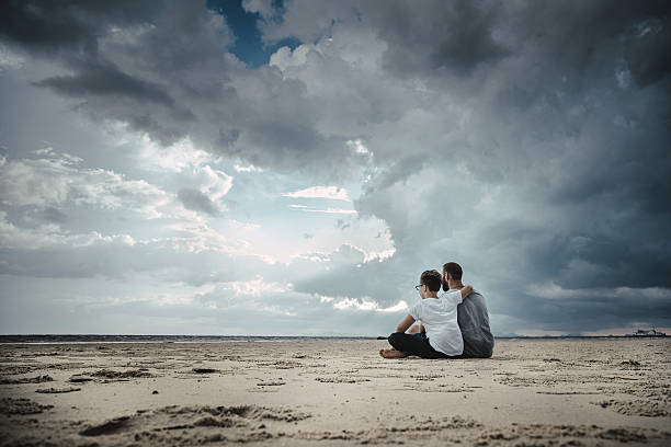 Father and son looking at sunset sitting on the beach. Stock photo.