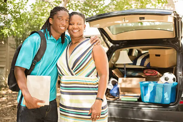 African descent boy heads off to college or moves away from home.  The 18-year-olds' mother is helping him pack up his car as he gets ready for the big move.  He is excited to start his college adventures. He wears a backpack and carries textbooks.  Family events.  Back to school.