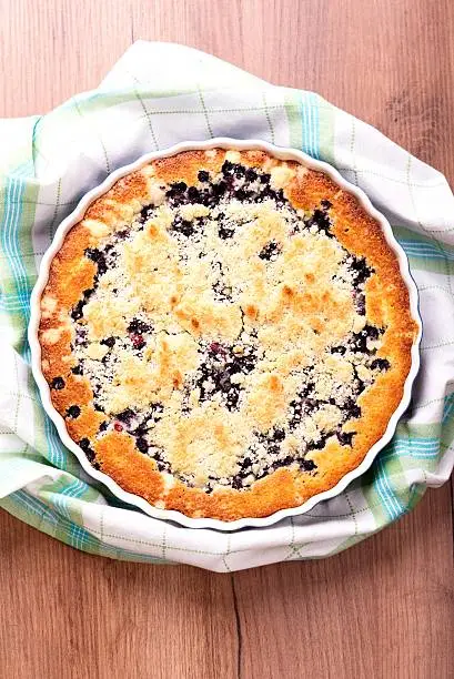 Vertical photo( (top view) of homemade blueberry pie which is made from wild berries harvested from forest. Cake is in blue baking dish placed on green checkered towel which is on brown wooden board.