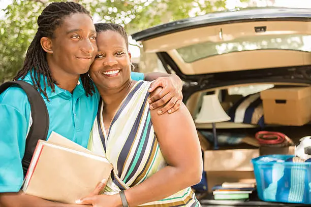 African descent boy heads off to college or moves away from home.  The 18-year-olds' mother is helping him pack up his car as he gets ready for the big move.  He is excited to start his college adventures and gives mom a big hug. He wears a backpack and carries textbooks.  Family events.  Back to school.