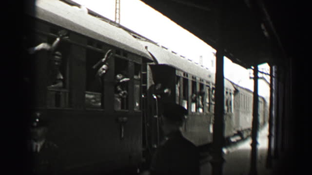 1937: Railroad leaving station passengers wave goodbye from train cars.