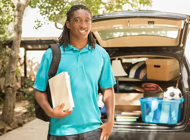 African descent boy heads off to college or moves away from home.  The 18-year-olds is packing up his car as he gets ready for the big move.  He is excited to start his college adventures. He wears a backpack and carries textbooks.   Back to school.