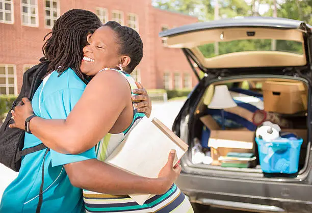 African descent boy heads off to college.  The 18-year-olds' mother is helping him unpack his car and says goodbye as he moves into the college campus dorm.  He is excited to start his school adventures. He  carries backpack and textbooks.  Family events.  Back to school.