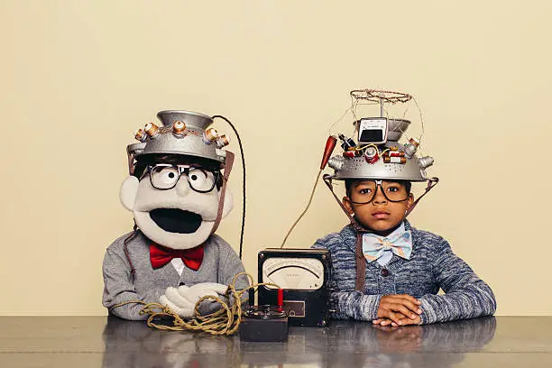 A nerd boy with friendly puppet are dressed in casual clothing, glasses and bow ties. They are experimenting with a homemade science project. They are both sitting at a table, with helmets on their heads in front of a beige background. Retro styling.
