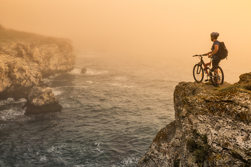 Mountainbiker standing on top of the rock and resting. Wears sportswear, helmet and backpack. Sea, waves and rocks on background.
