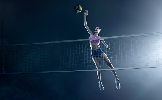 Beautiful female volleyball player performs an emotional game moment on the dark foggy background with net while holding a ball. She is wearing an unbranded sports cloth.