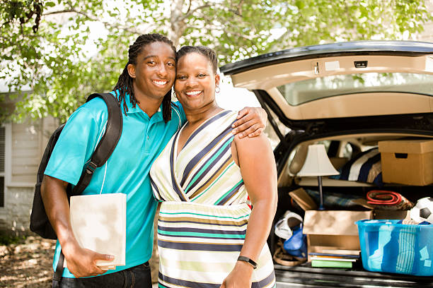 African-descent boy moves off to college.  Packed car, mom. Home. African descent boy heads off to college or moves away from home.  The 18-year-olds' mother is helping him pack up his car as he gets ready for the big move.  He is excited to start his college adventures. He wears a backpack and carries textbooks.  Family events.  Back to school. college student and parent stock pictures, royalty-free photos & images