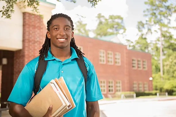 Back to school.   Handsome, African descent boy heads off to college.  The 18-year-old is excited to start his first day of school.  He carries a backpack and textbooks.  College building in background.