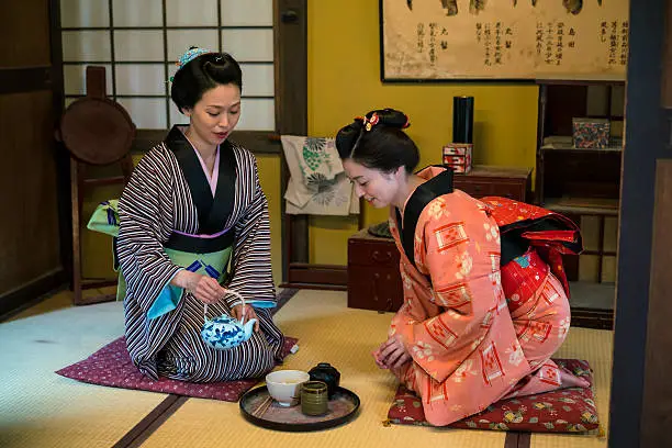 Vview of two women in traditional kimono, kneeling on tatami having  cup of tea which is in front of them on tatami. They are in traditional Japanese old house. This is in Toei studios in Kyoto with old buildings from Samurai times.