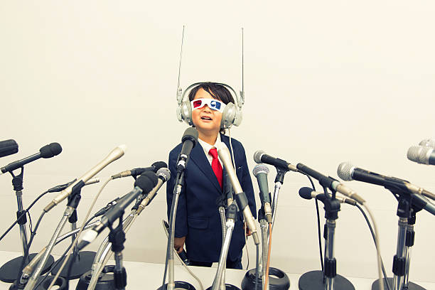 A young Japanese boy in business suit, homemade communication headset and 3D glasses stands in front of a large number of microphones. He is hoping for the best communication and public relations for his business. He is smiling at the camera.  