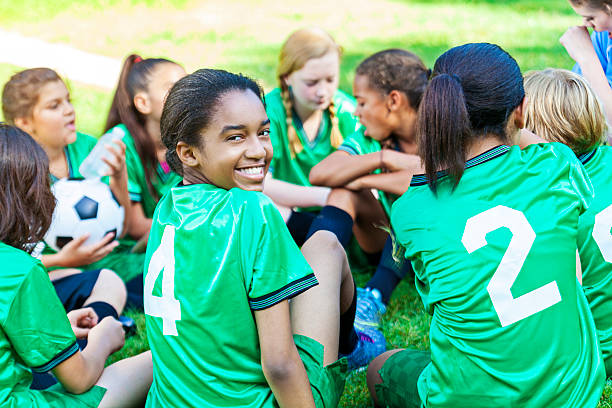 Beautiful African American girl smiling with her soccer team Beautiful African American girl turning around and smiling at camera while sitting down with her all girls soccer team. Their uniforms are a bright green. sports activity stock pictures, royalty-free photos & images