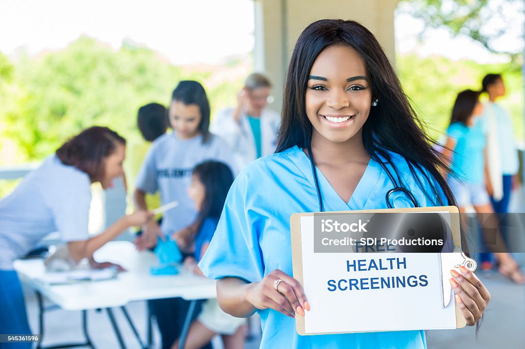 Beautiful African American Woman holding a ""Free Health Screenings"" sign Beautiful African American nurse holding a "Free Health Screenings" sign. She is wearing light blue scrubs. Her hair is long straight and black. She has a stethoscope around her neck. There are volunteers and patients working behind her. Film and Television Screening Stock Photo