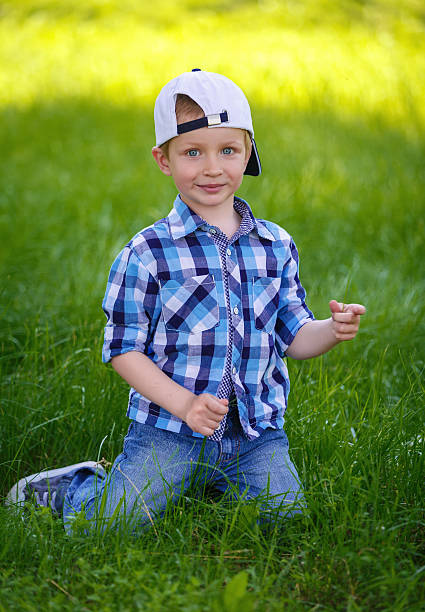 Little boy in a blue plaid shirt is sitting on the green grass in the park