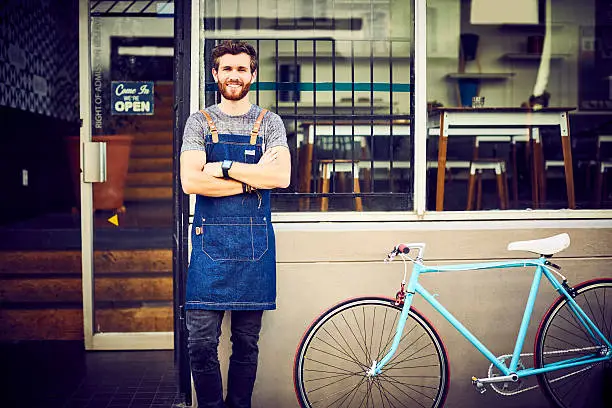 Photo of Confident barista smiling by bicycle outside cafe