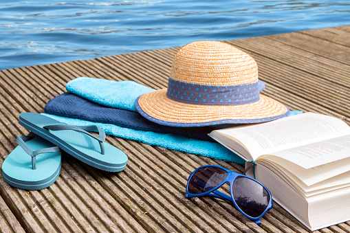 summer vacation, blue water and accessories for beach holidays as straw hat, flip flops,  sunglasses, turquoise towels and a book on a wooden bathing pier at the pool, selected focus and narrow depth of field