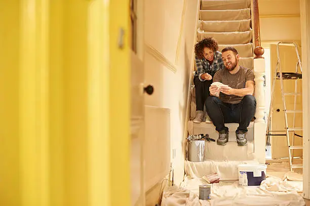 A young couple doing up their hallway in the old house they have bought . They are sitting on the dust sheets on the stairs and looking through the paint sample cards choosing the final colour for the beige hall .