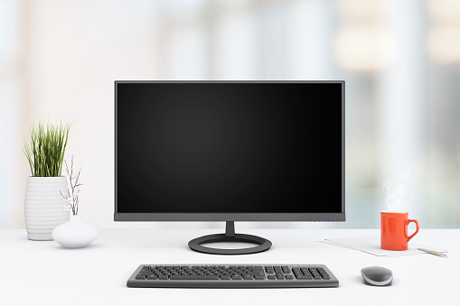 View at a working desk,  with a large PC monitor,lamp, a plant, coffee cup, with a blurry out of focus business background.