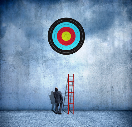 A businessman stands with his back to the camera and his hands in his pockets as he leans back and looks up at the bulls eye on a target on a wall high above him.  A ladder that is too short leans against the wall next to him.