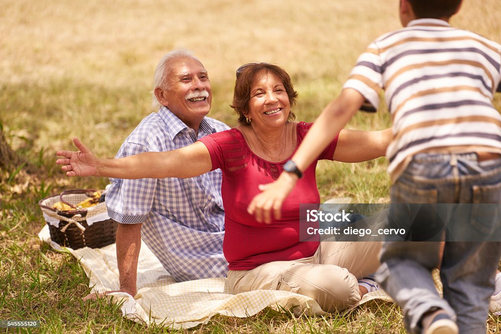 Grandparents Senior Couple Hugging Young Boy At Picnic Old people, senior couple, elderly man and woman. Outdoor family having fun with happy grandpa and grandma hugging boy at picnic in park. Latin American and Hispanic Ethnicity Stock Photo