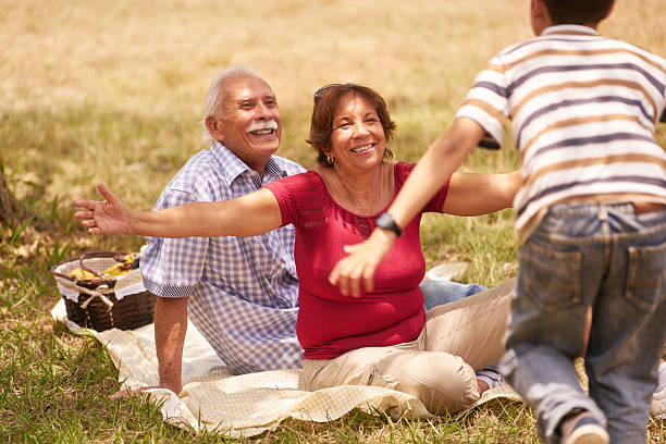 Grandparents Senior Couple Hugging Young Boy At Picnic Old people, senior couple, elderly man and woman. Outdoor family having fun with happy grandpa and grandma hugging boy at picnic in park. hispanic grandmother stock pictures, royalty-free photos & images