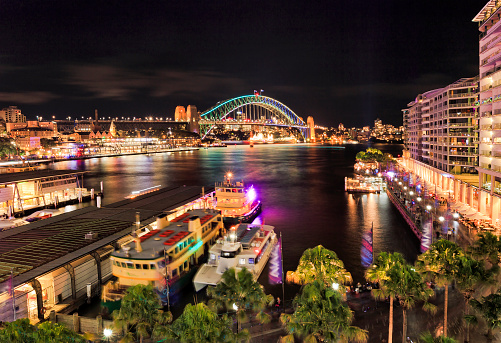 Elevated panorama of Sydney harbour circular quay bay at night during bright illumination by light towards the arch of Harbour bridge.