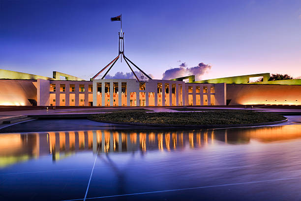 CAN Parliament Facade water ligh set Australian national parliament house in Canberra. Facade of the buidling brightly illuminated and reflecting in blurred water of fountain pond under waving national flag on flagpole at sunset. canberra photos stock pictures, royalty-free photos & images
