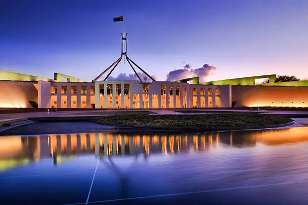 Australian national parliament house in Canberra. Facade of the buidling brightly illuminated and reflecting in blurred water of fountain pond under waving national flag on flagpole at sunset.