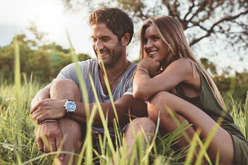 Portrait of smiling young couple in love sitting together outdoors in field. Happy young man and woman in meadow.