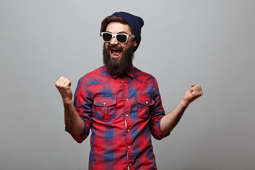 happy hipster man with beard wearing red checkered shirt and sunglasses exults isolated on grey background