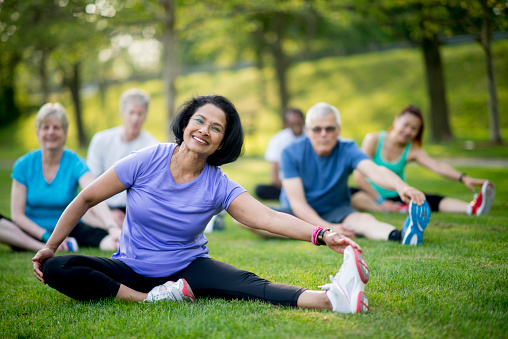 A multi-ethnic group of senior adults are taking an outdoor yoga class at the park. They are sitting in the grass and stretching to increase flexibility and reduce injury.
