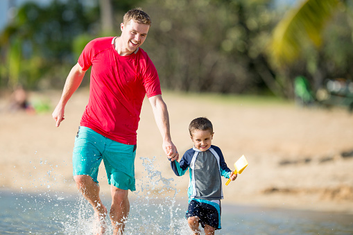 A father and son are running through the water next to the beach while on vacation in Hawaii. They are smiling and laughing together.