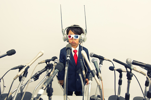 A young Japanese boy in business suit, homemade communication headset and 3D glasses stands in front of a large number of microphones. He has a blank face and is nervous as he is speaking out for his business. 