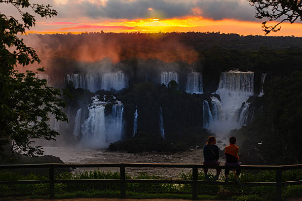 Iguacu, Brazil: Silhouette Of Tourists At The Famous Falls, Watching A Dramatic Sunset Over The Falls Near The Devil’s Throat Iguacu, Brazil - November 06, 2010: A couple of lady tourists watch the sunset over the Iguassu falls, near the Devil's Throat, in Brazil, South America. Located between the countries of Brazil and Argentina, it is considered one of the new seven natural wonder of the world. An amazing volume of water goes over the falls at so many adjacent locations, making it one the most spectacular waterfalls in the world. Birds and sections of the rainforest complete the photograph. Shot at sunset time; horizontal format. Copy space. devils throat stock pictures, royalty-free photos & images