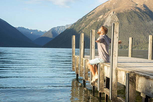 istock Young man sits on jetty above lake, arms outstretched 545675190