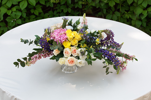 Beautiful flower composition outdoors. Wedding floristic decoration at white round table, bouquet of colorful wildflowers and white roses in glass vase at green leaves background