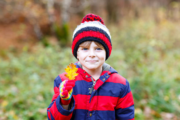 Portrait of happy cute little kid boy with autumn leaves background in colorful clothing. Funny child having fun in fall forest or park on cold day. With hat and gloves
