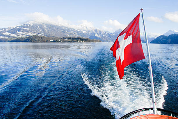 A close view of a vintage boat cruising on a vibrant turquoise alpine lake, proudly displaying the Swiss flag with a dramatic mountainous skyline in the background.
