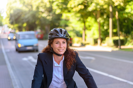 Attractive professional woman wearing a safety helmet cycling to work along an urban street with traffic, close up upper body view
