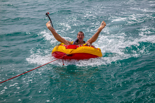Man sitting in inflatable ring towed by a boat in the water and making faces to the camera and holding Go Pro camera.