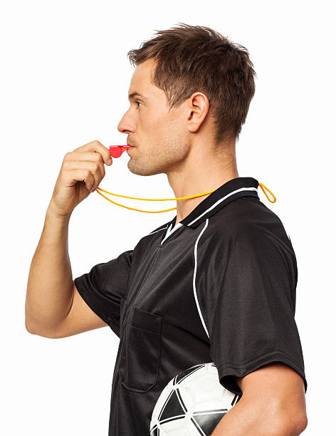 Side View Of Referee Whistling While Holding Soccer Ball Side view of referee whistling while holding soccer ball under arm against white background. Horizontal shot. whistling stock pictures, royalty-free photos & images