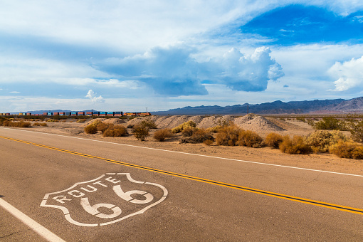 U.S. Route 66 highway, with sign on asphalt and a long train in the background, near amboy, california. Located in the mojave dessert. Photo made on a motorcycle roadtrip.