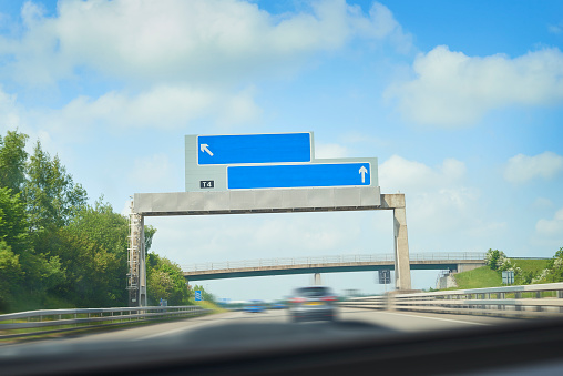 A car is approaching a motorway information sign. The sign has had names removed for people to add in the places required.  This sign is from a uk motorway. The image is taken from inside a car with the very top of the dashboard visible. All lanes of the motorway are visible and other traffic in the distance. ( vehicles have been heavily retouched and blurred to be made unrecognisable )