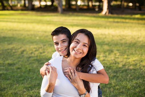 A teen latin boy embracing his mother from behind, holding hands and smiling at the camera in a horizontal waist up shot outdoors.